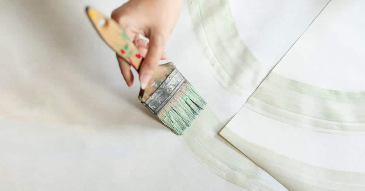 Une main tenant un pinceau applique une couche de peinture sur un mur nu d'un côté et recouvert de papier peint de l'autre.